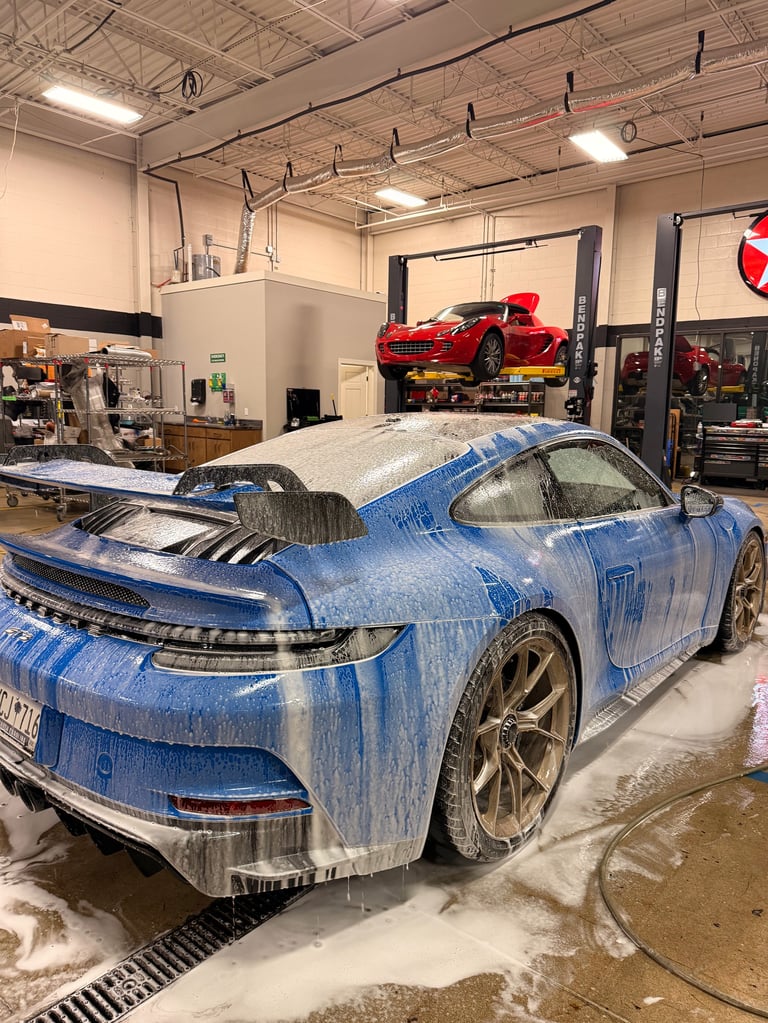 Blue sports car covered in soap suds being washed in an auto shop with red vehicle on lift in background