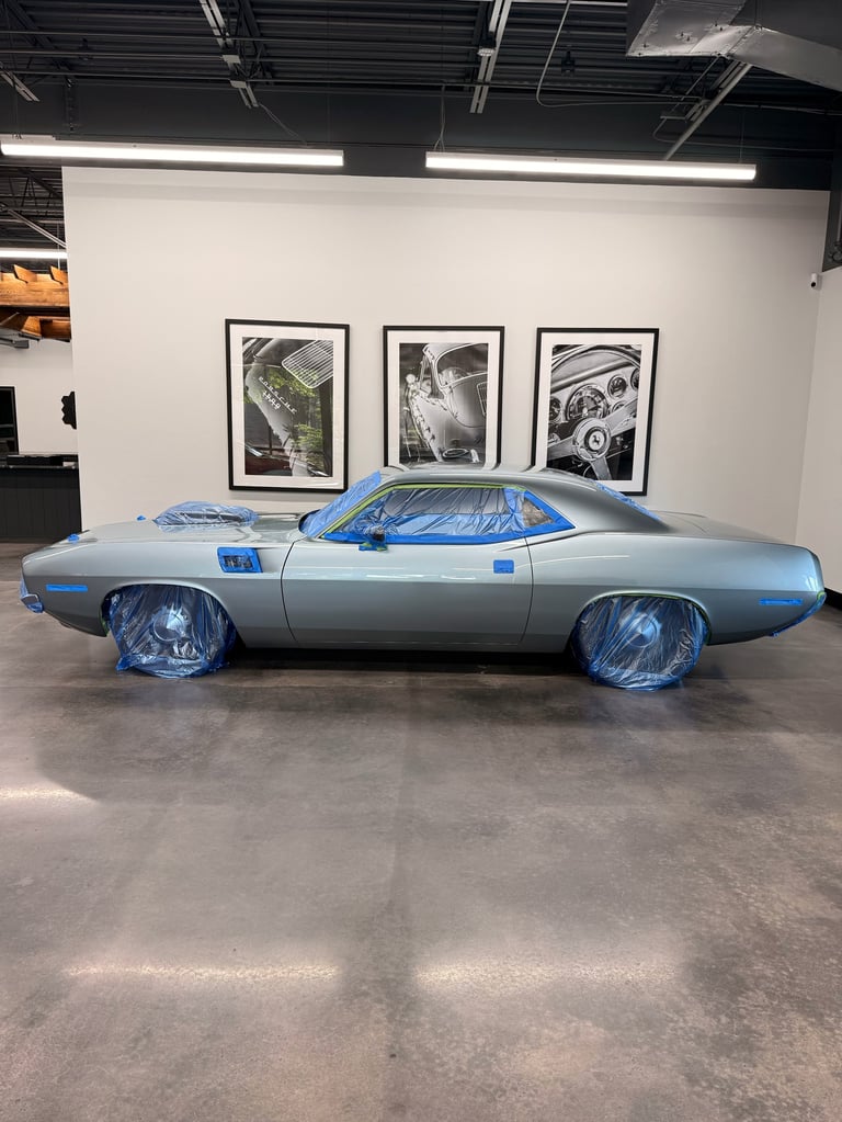 Silver classic muscle car with blue accents displayed in a gallery with black-framed photographs on white wall and industrial ceiling