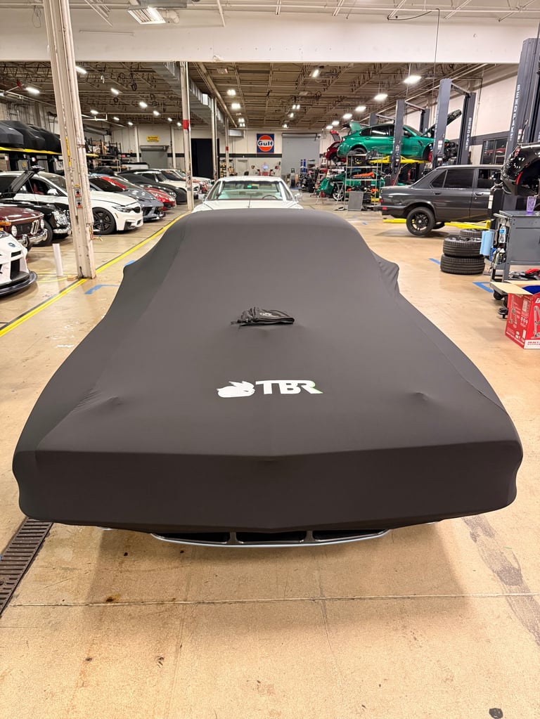 Car covered with black TBR car cover in auto repair shop with other vehicles visible in background