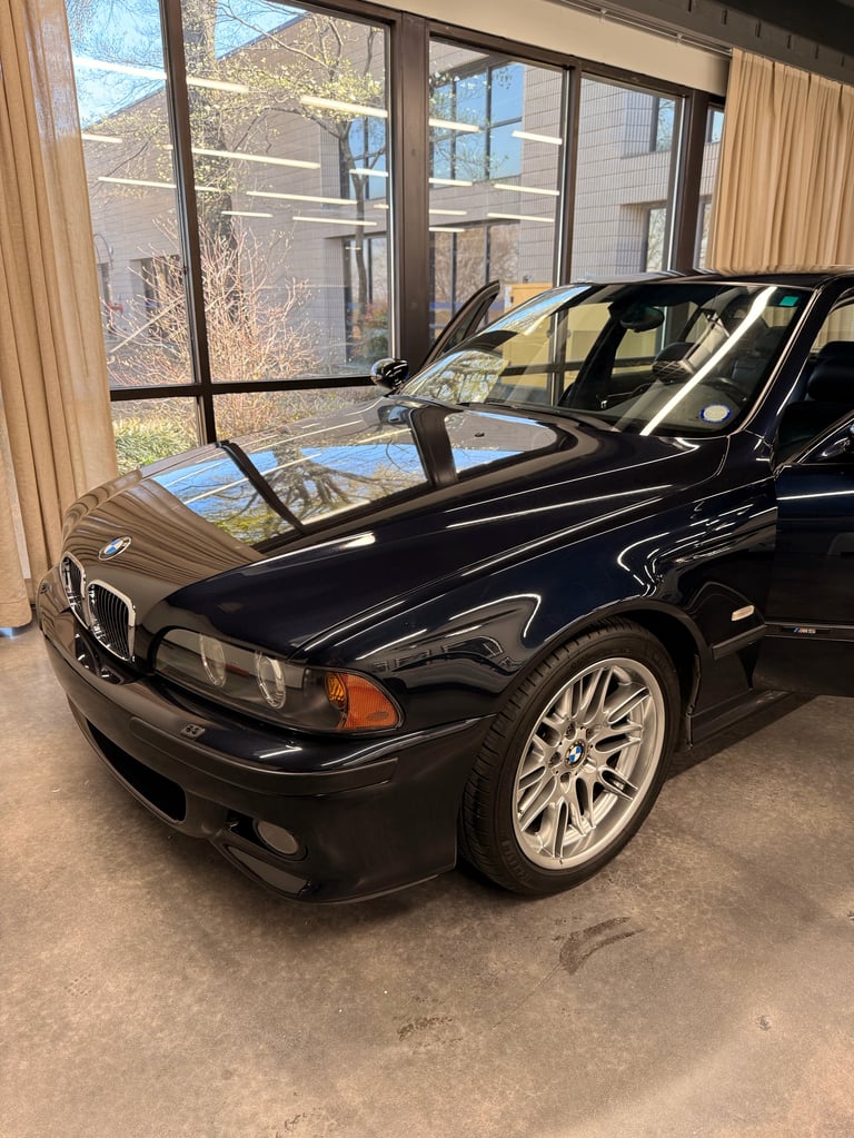 Black BMW M5 sedan displayed indoors with hood open, showing engine, in a showroom with large windows