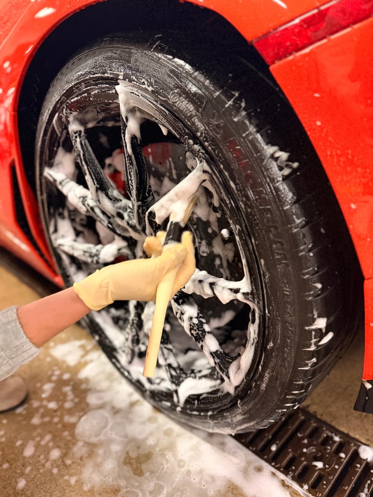 Hand in yellow glove washing a black car wheel with white soapy water on a red vehicle