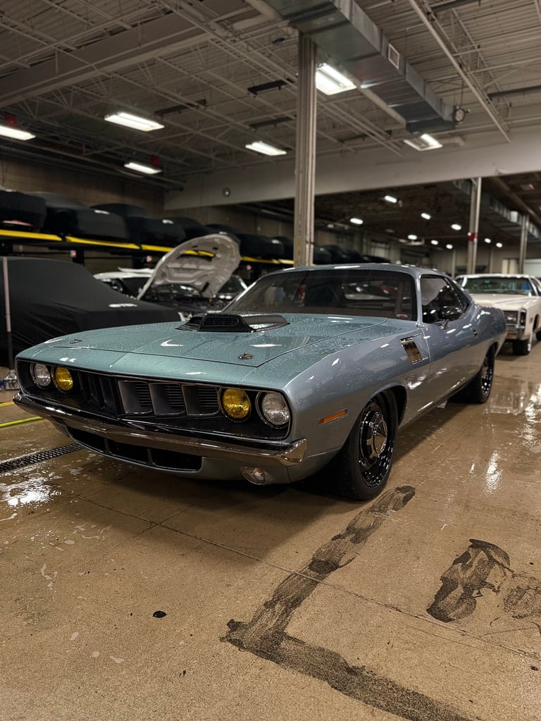 Light blue 1970s Plymouth muscle car with hood open displayed in warehouse with other vehicles