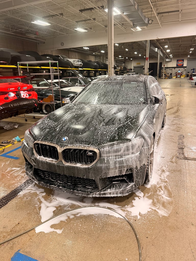 BMW car covered in white foam during wash in an indoor garage with racing vehicles and equipment nearby