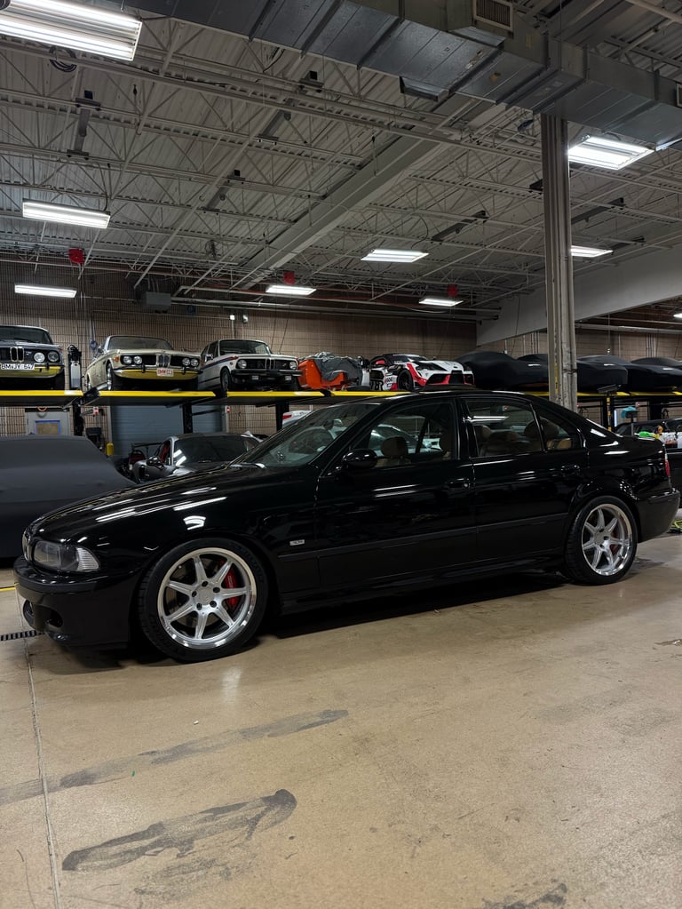 Black luxury sedan with silver wheels displayed in an industrial warehouse garage with fluorescent lighting and other vehicles in background