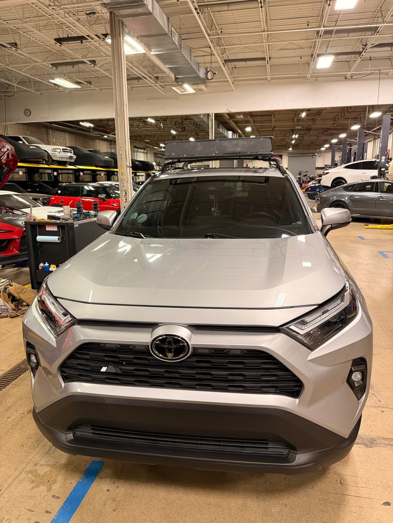 White Toyota RAV4 SUV displayed in a car dealership showroom with roof rack and other vehicles visible in background