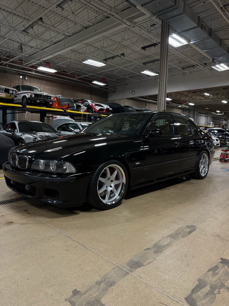 Black BMW M5 sedan with custom wheels parked in an industrial warehouse with other vehicles visible in the background