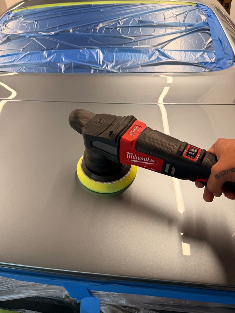 Person holding a red and black Milwaukee orbital polisher with yellow foam pad on a white car hood with blue protective material visible
