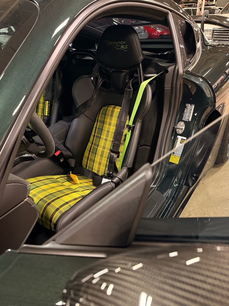 Car interior showing black racing seat with yellow and black checkered pattern and neon yellow harness