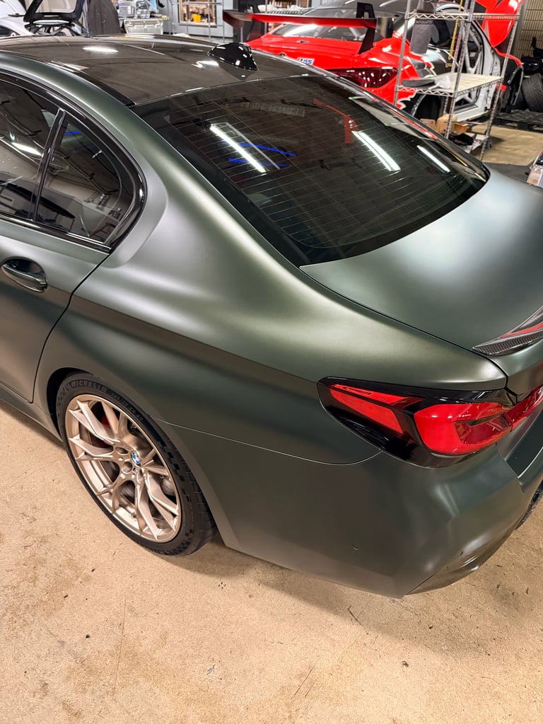 Silver sedan with matte finish parked in workshop, red race car visible in background