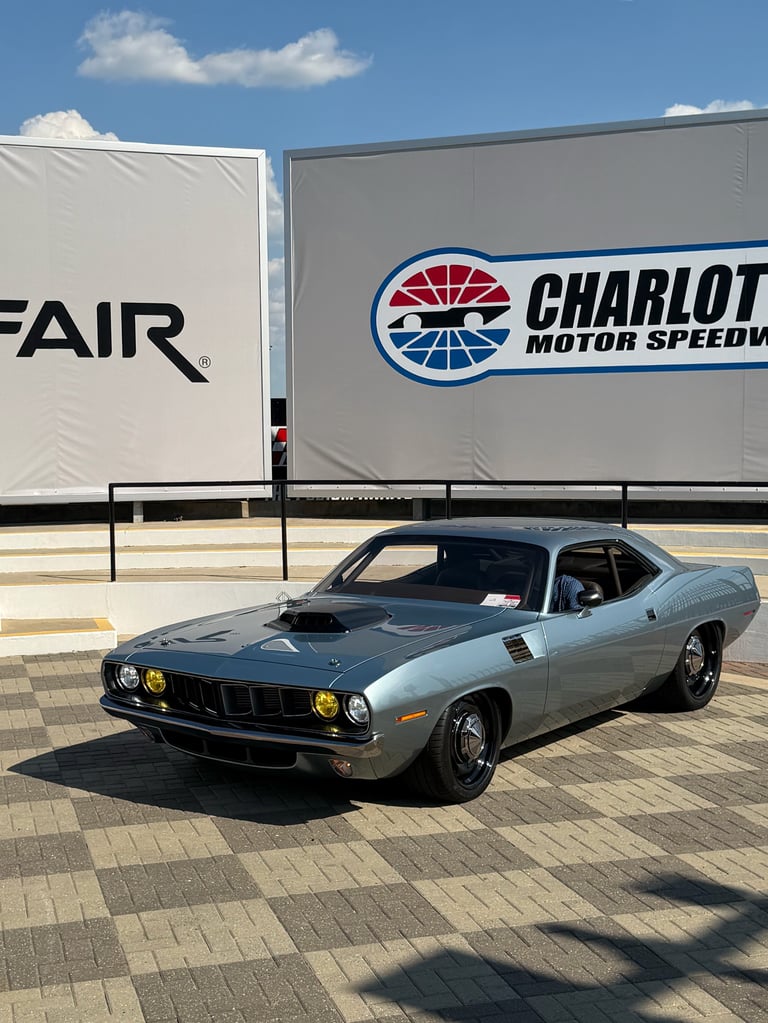 Light blue vintage muscle car parked in front of Charlotte Motor Speedway banner at fairground