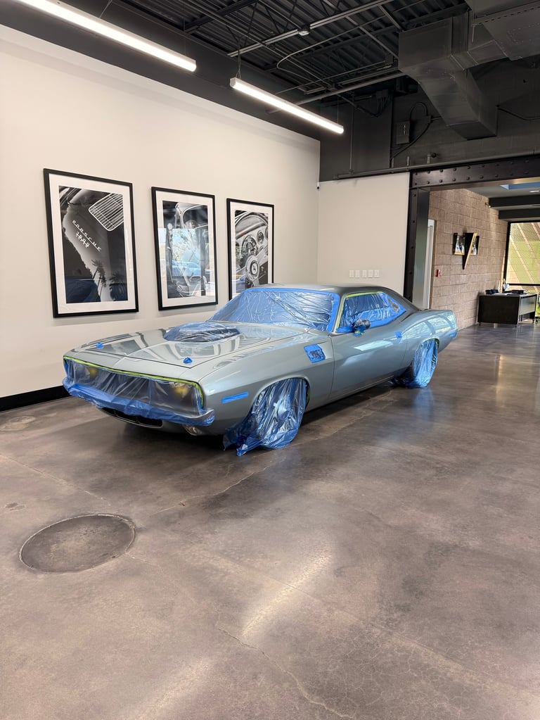 Blue and silver muscle car wrapped in plastic displayed in an industrial gallery space with framed photographs on the wall
