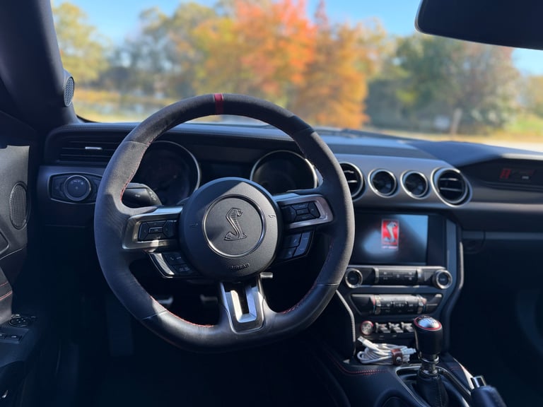 Interior of a Ford Mustang sports car with dark steering wheel and dashboard, autumn foliage visible through windshield