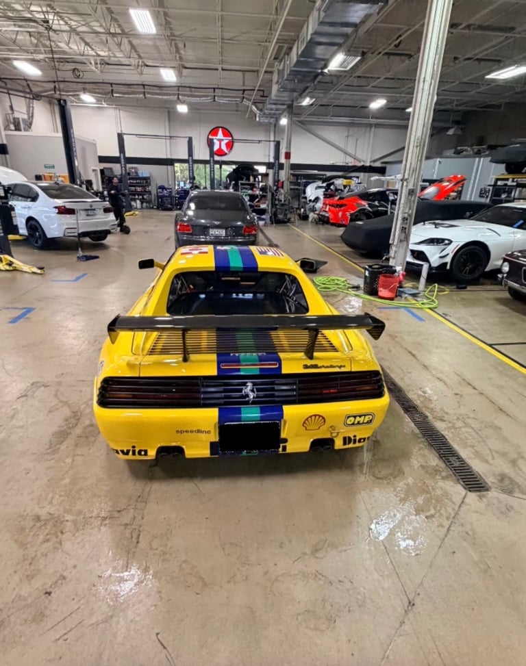 Yellow sports car with blue racing stripes in a garage workshop with other vehicles and equipment visible