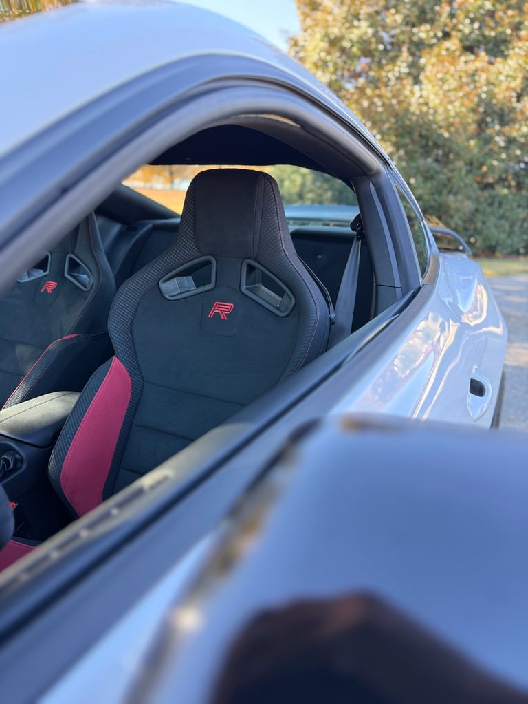 View through car window of black and red sports car seat with R logo, showing interior and outdoor background