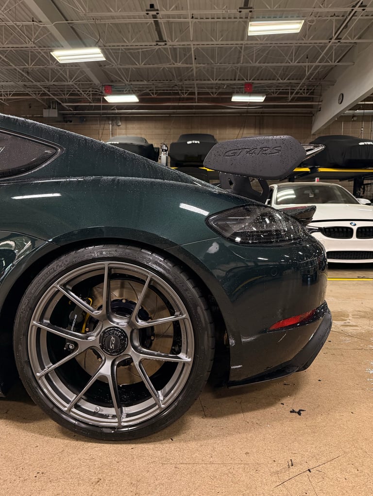 Dark green sports car with metallic wheels in an indoor warehouse garage, other vehicles visible in background