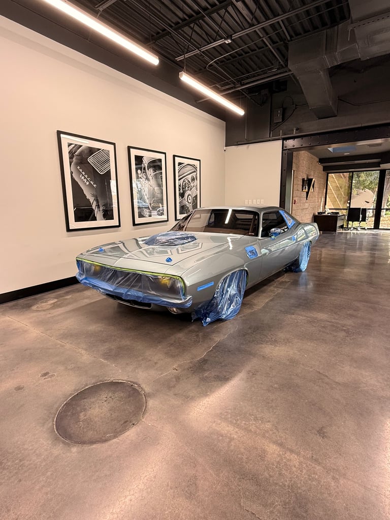 Classic blue car with chrome wheels displayed in modern gallery space with industrial ceiling and framed artwork on white walls